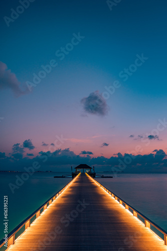 Amazing sunset panoramic beach Maldives. Luxury resort villas long wooden pier seascape with soft led lights under colorful sky view with calm sea and relaxing tropical mood. Amazing travel landscape