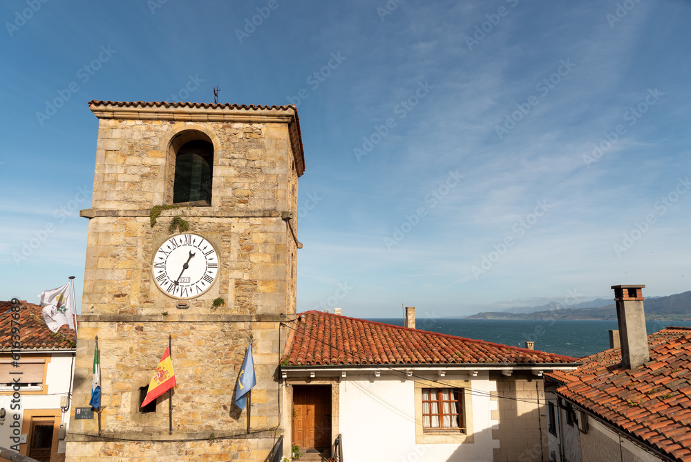 Detail plan of the clock tower of the town hall of the touristic coastal town of Lastres with its medieval stone architecture and flags flying on its façade on a sunny day.