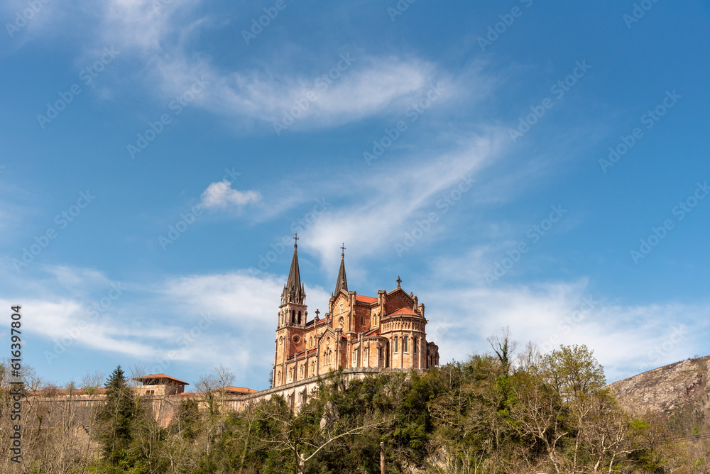 Impressive panoramic view of the basilica of Santa María la Real de Covadonga in the touristic province of Asturias on a hill with its reddish medieval architecture surrounded by greenery.