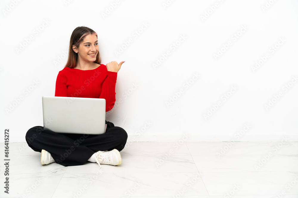 Young caucasian woman with a laptop sitting on the floor pointing to the side to present a product Young caucasian woman with a laptop sitting on the floor pointing to the side to present a product