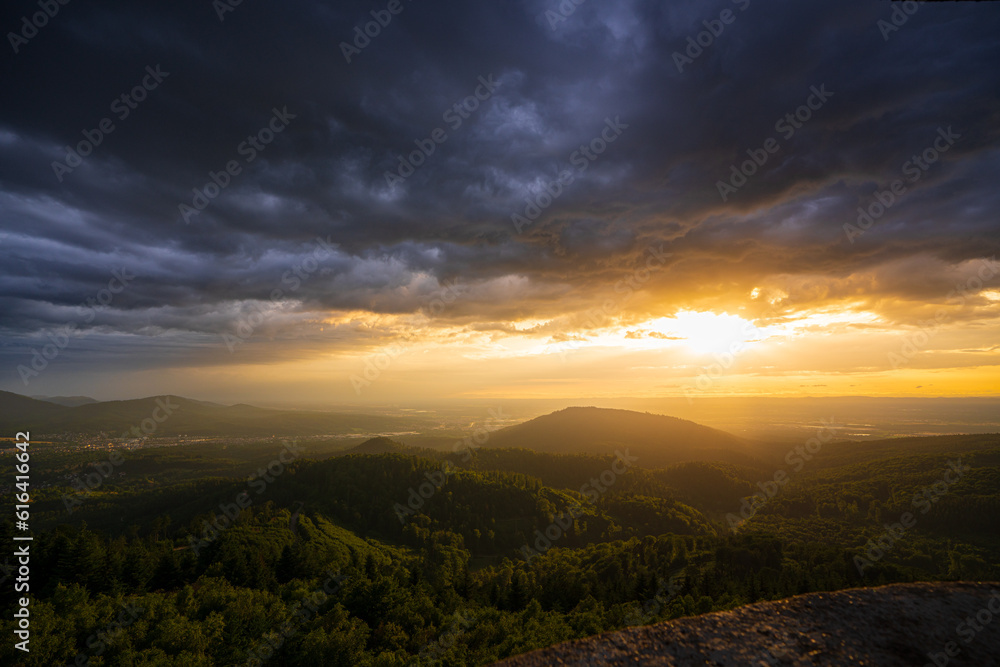 A threatening storm cloud passes over the Murg valley in the northern Black Forest while in the background the setting sun shines in a pleasantly warm light.