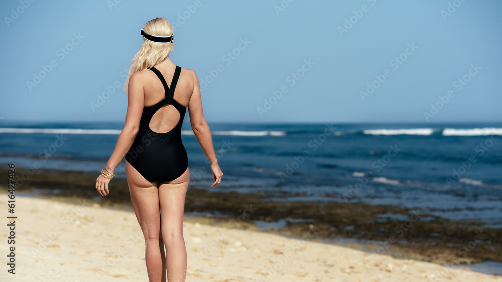 A girl in a black swimsuit stands on the beach and looks at the ocean. View of a woman's back against the background of water and sand. Beach holidays in Bali. A blonde with tanned skin enjoys. A girl in a black swimsuit stands on the beach and looks at the ocean. View of a woman's back against the background of water and sand. Beach holidays in Bali. A blonde with tanned skin enjoys.