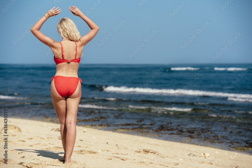 A red in a purple swimsuit stands on the beach and looks at the ocean. View of a woman's back against the background of water and sand. Beach holidays in Bali. A blonde with tanned skin enjoys. A red in a purple swimsuit stands on the beach and looks at the ocean. View of a woman's back against the background of water and sand. Beach holidays in Bali. A blonde with tanned skin enjoys.