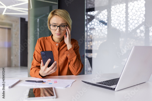 Fototapeta Sad upset woman pensive with phone in hands working inside office at workplace, businesswoman financier received notification message with bad news online, uses app on smartphone