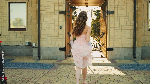 Fantasy woman running down vintage old style city street to gate road way paving stones. Princess girl pink long vintage dress fabric waving. back rear view queen. Summer day outdoor sun divine light
