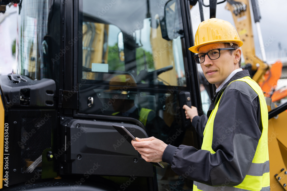 Worker in a helmet with a digital tablet on the background of construction machine.. Worker in a helmet with a digital tablet on the background of construction machine..