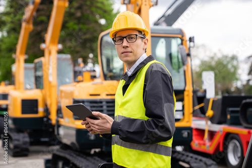 Engineer in a helmet with a digital tablet stands next to construction excavators.