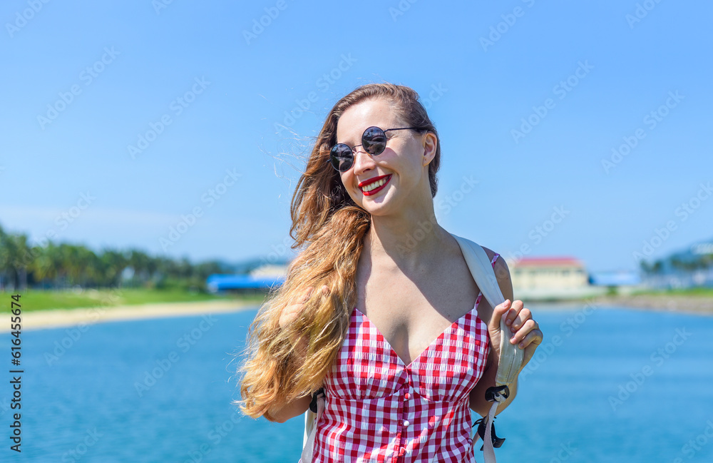 Bright blond young woman standing with backpack near sea in summer 