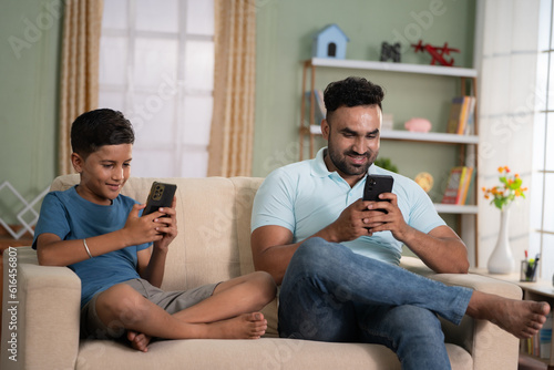 focus on father, Indian father and son busy on there mobile phones while sitting on sofa at home - concept of technology addiction, social media and togetherness