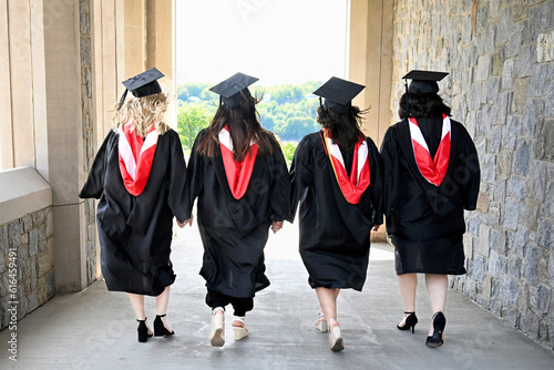 Lifestyle portrait from back of group of female college graduates holding hands walking in a row wearing caps and gowns