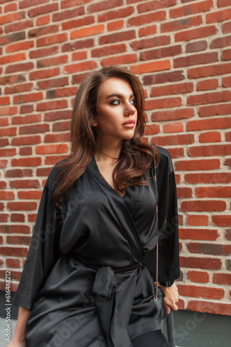 Fashionable beautiful young chic woman model in elegant black suit sitting near brick vintage wall