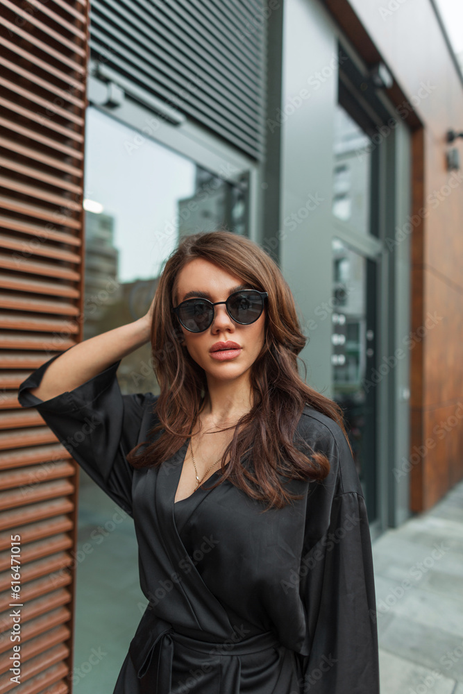 Fashion beautiful glamor chic woman model with vintage cool sunglasses in trendy elegant black clothes walking on the street near the building
