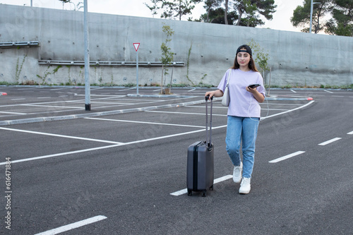 Girl with a suitcase talking on a mobile phone while standing in a parking lot. The student went to study. University campus. Day light. Travel, education concept. Lifestyle. 