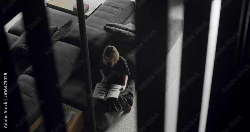 A woman is sitting on the couch and reading a book. Modern contemporary Minimalist living room with white wall and black furniture. Luxury Modern House Interior With Corner Sofa and Bookshelf. A woman is sitting on the couch and reading a book. Modern contemporary Minimalist living room with white wall and black furniture. Luxury Modern House Interior With Corner Sofa and Bookshelf.