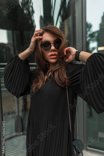 Urban female fashion portrait of stylish beautiful fashionable woman with cool vintage black sunglasses in trendy dress with small bag walks and poses on the street.
