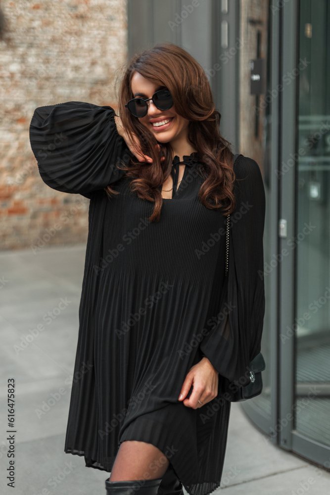 Happy beautiful glamor woman model with hairstyle with stylish vintage sunglasses in a fashion black dress with a handbag walks and has fun in the city. Pretty lady