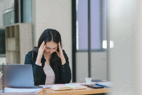 The stressed and exhausted millennial Asian businesswoman is seen sitting at her office desk with her hand on her head, indicating a hard working day where she is overloaded with work.