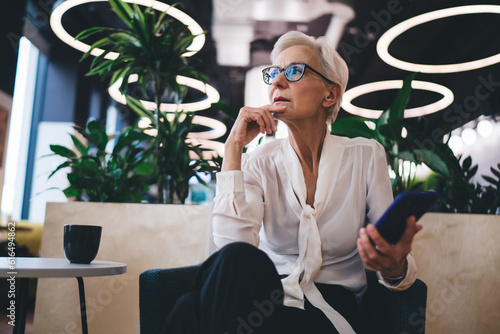 Thoughtful aged woman sitting with smartphone in modern office