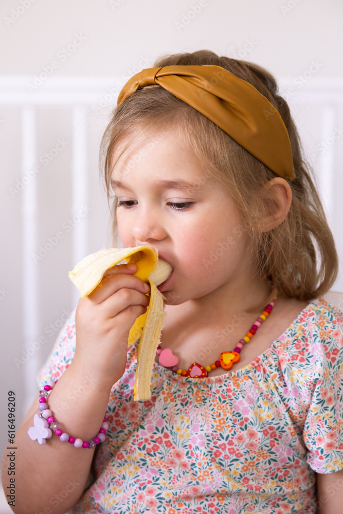 Pretty five-year old little girl with headband in her fine blond hair, a cute necklace and dress sitting eating a banana Pretty five-year old little girl with headband in her fine blond hair, a cute necklace and dress sitting eating a banana