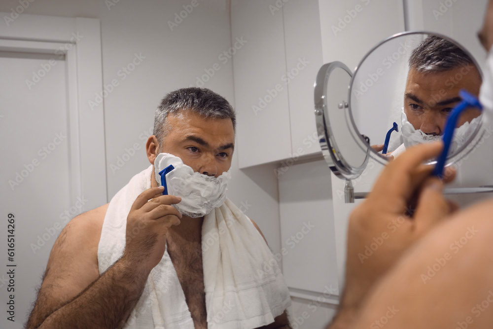 middle age man shaving in the bathroom in the morning, daily routine. serious man applying shaving foam at bathroom mirror. male shaving beard with razor blade at bathroom