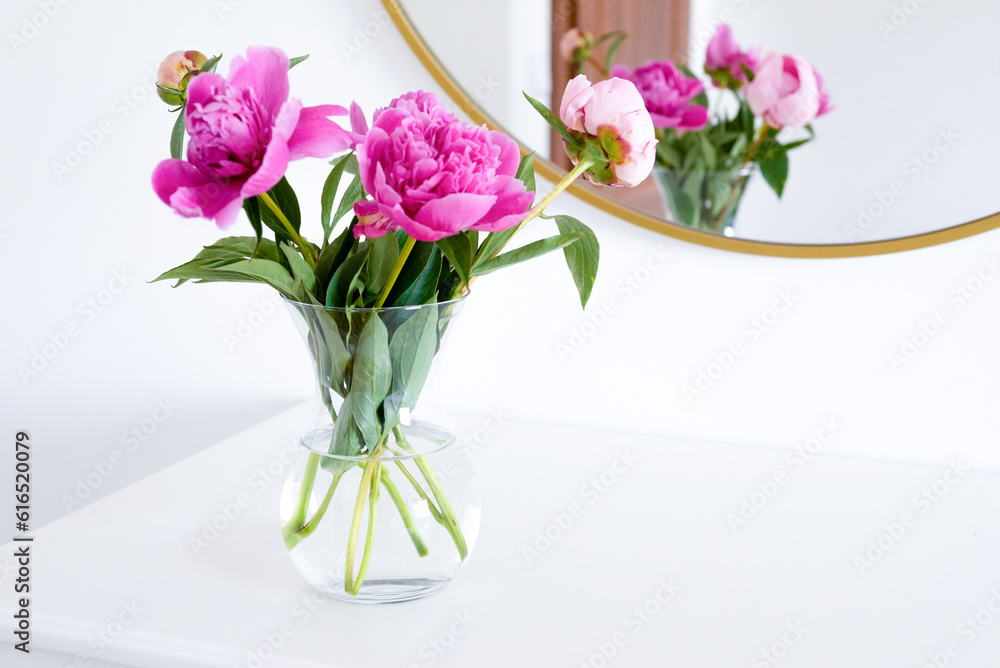 Fototapeta premium Pink peonies in the apartment. Home decoration, pink peonies on table in white room. Vase with beautiful peony flowers on table in bedroom. Flowers in a vase at home. Mirror reflection, gold frame