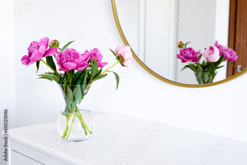 Pink peonies in the apartment. Home decoration, pink peonies on table in white room. Vase with beautiful peony flowers on table in bedroom. Flowers in a vase at home. Mirror reflection, gold frame