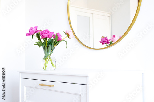 Pink peonies in the apartment. Home decoration, pink peonies on table in white room. Vase with beautiful peony flowers on table in bedroom. Flowers in a vase at home. Mirror reflection, gold frame