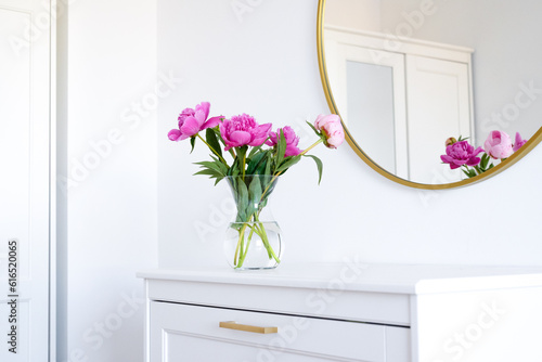 Pink peonies in the apartment. Home decoration, pink peonies on table in white room. Vase with beautiful peony flowers on table in bedroom. Flowers in a vase at home. Mirror reflection, gold frame