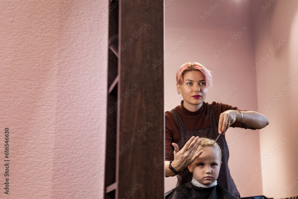 Hairdresser hands makes hairstyle for little blonde girl at mirror in barber shop. Hairstylist woman braiding braids for cute kid in barbershop, hairsalon. Haircare, beauty concept. Copy ad text space