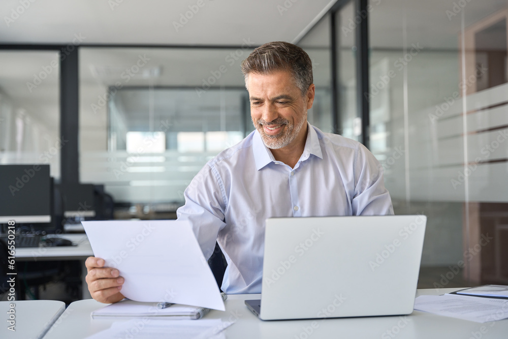Smiling mature professional business man manager looking at report in office. Happy mid aged businessman company executive working in office with laptop reading financial corporate account documents. Smiling mature professional business man manager looking at report in office. Happy mid aged businessman company executive working in office with laptop reading financial corporate account documents.