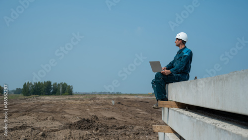 Caucasian male builder in hardhat sits on floor slabs and uses laptop at construction site. 