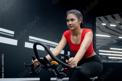 Fototapeta Naklejka Na Ścianę i Meble -  Young asian woman riding bicycle machine for workout training in fitness gym club, female cycling for exercise with motivation and concentration for wellbeing and weightloss, sport and healthcare.