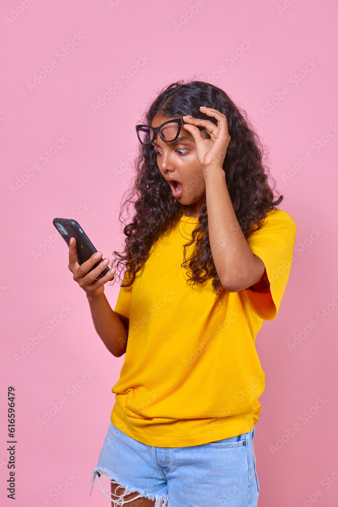 Young surprised Indian woman teenager with mobile phone in hands opens mouth wide and raises glasses sees shocking news on website or in received email message stands on isolated pink background. Young surprised Indian woman teenager with mobile phone in hands opens mouth wide and raises glasses sees shocking news on website or in received email message stands on isolated pink background.