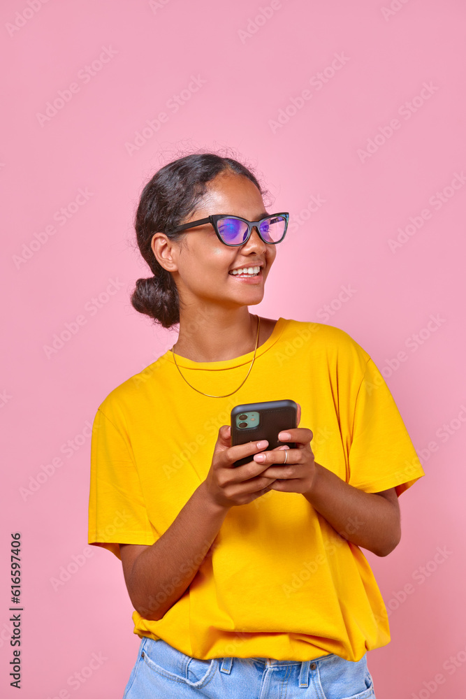 Fototapeta premium Young positive ethnic Indian woman teenager in casual clothes holding phone and laughing chatting with friend from school or arranging walk together on weekend stands on pink background.
