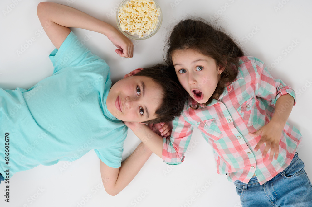 Happy children with popcorn bowl smile at camera, express surprise and amazement, lying on their backs on isolated white studio background. Kids. Childhood. Entertainment. Lifestyle. Leisure activity Happy children with popcorn bowl smile at camera, express surprise and amazement, lying on their backs on isolated white studio background. Kids. Childhood. Entertainment. Lifestyle. Leisure activity