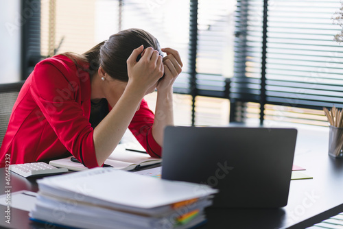 The stressed and exhausted millennial businesswoman is seen sitting at her office desk with her hand on her head, indicating a hard working day where she is overloaded with work.