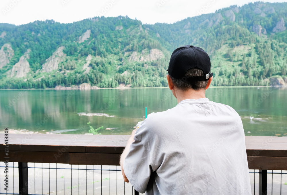 Back view of male traveler sitting on wooden bench while enjoying amazing nature in mountains. Man drinking coffee outdoor in morning.