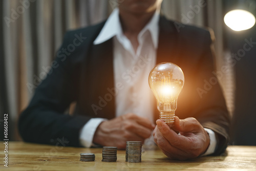 Businessman holding a light bulb Concept of saving money with hands putting coins on wooden table floor