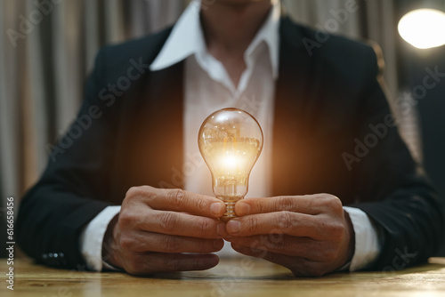 Businessman holding a light bulb Concept of saving money with hands putting coins on wooden table floor