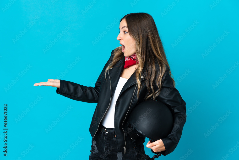 Romanian woman holding a motorcycle helmet isolated on blue background with surprise expression while looking side Romanian woman holding a motorcycle helmet isolated on blue background with surprise expression while looking side