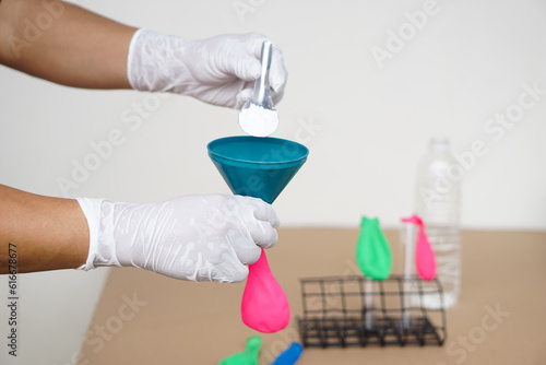 Closeup hands wears gloves holds funnel and flat pink balloon, put spoon of baking soda powder to pour into balloon. Concept, science experiment.             © Sanhanat