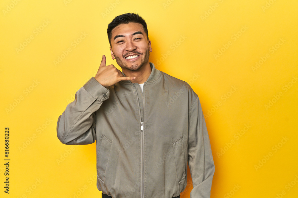 Stylish Asian man in gray jacket on yellow studio showing a mobile phone call gesture with fingers. Stylish Asian man in gray jacket on yellow studio showing a mobile phone call gesture with fingers.