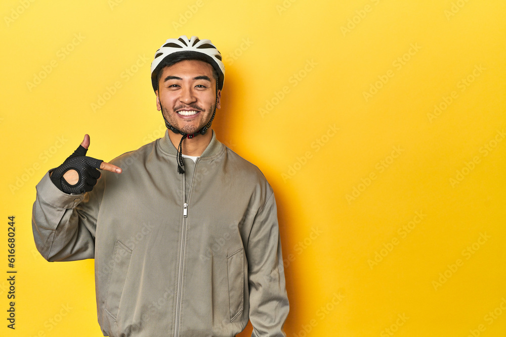 Asian man in bike helmet and gloves, yellow studio backdrop person pointing by hand to a shirt copy space, proud and confident Asian man in bike helmet and gloves, yellow studio backdrop person pointing by hand to a shirt copy space, proud and confident