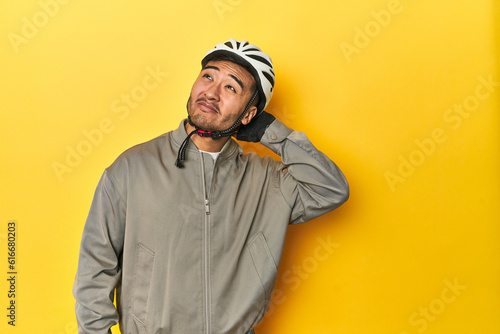 Asian man in bike helmet and gloves, yellow studio backdrop touching back of head, thinking and making a choice. © Asier