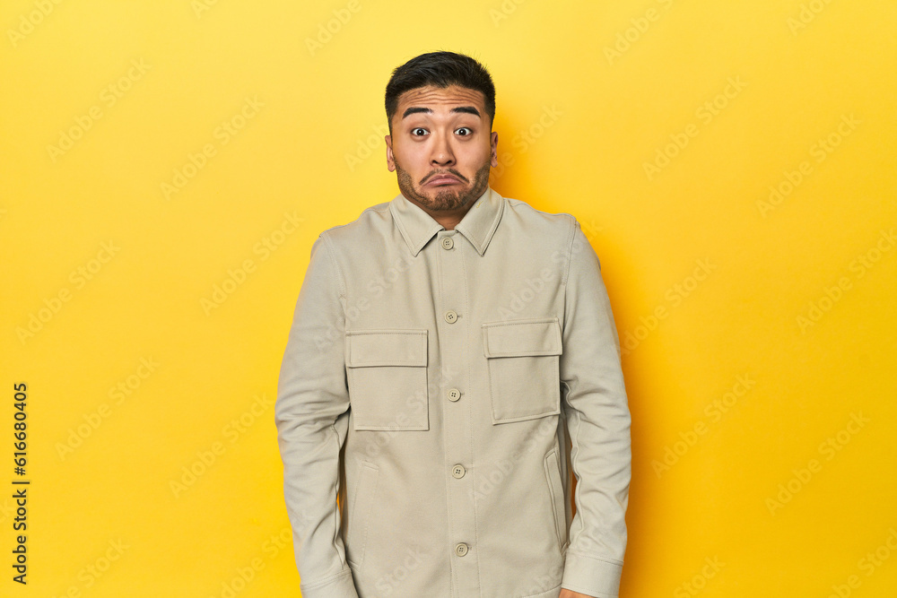 Asian man in stylish gray shirt on yellow studio shrugs shoulders and open eyes confused.