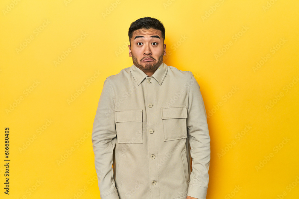 Fototapeta premium Asian man in stylish gray shirt on yellow studio shrugs shoulders and open eyes confused.