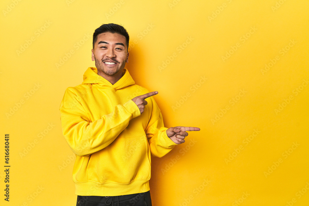 Bold Asian man in yellow hoodie on yellow studio excited pointing with forefingers away. Bold Asian man in yellow hoodie on yellow studio excited pointing with forefingers away.