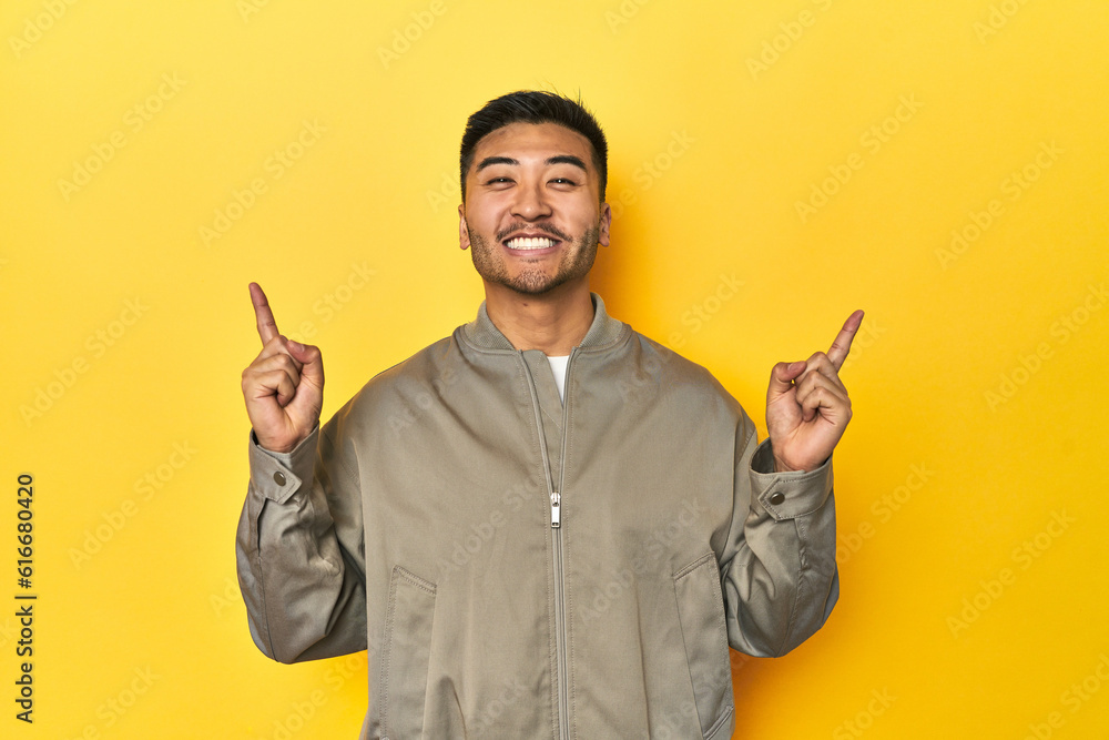 Stylish Asian man in gray jacket on yellow studio indicates with both fore fingers up showing a blank space.