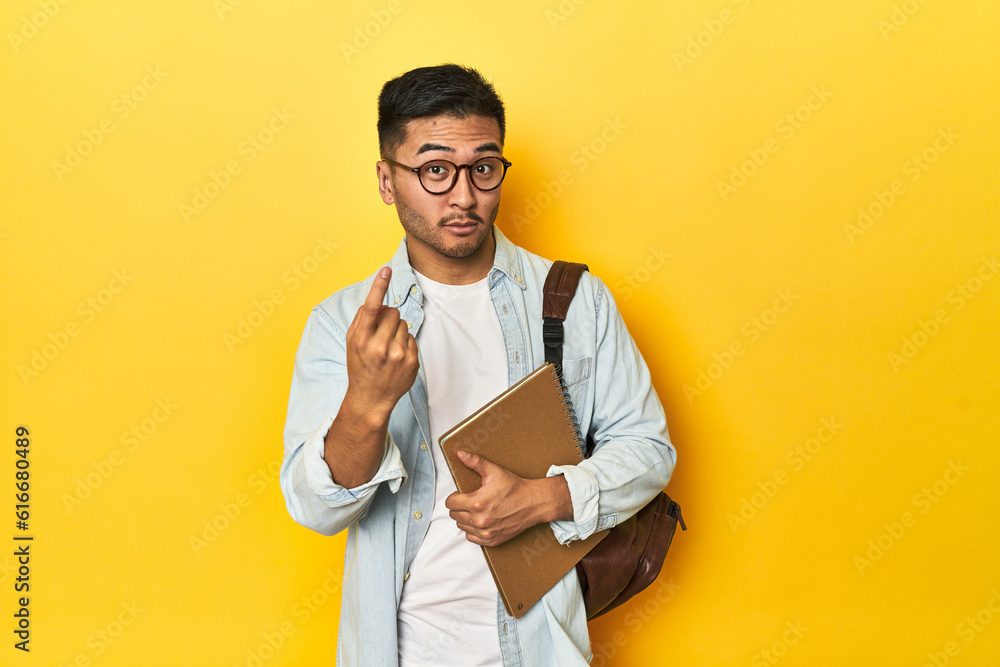 Asian student with backpack and notebook, yellow studio backdrop pointing with finger at you as if inviting come closer. Asian student with backpack and notebook, yellow studio backdrop pointing with finger at you as if inviting come closer.
