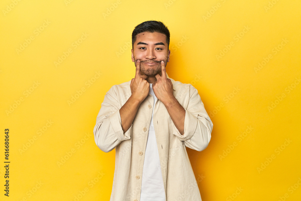 Casual Asian man with open shirt, white tee on yellow studio doubting between two options.